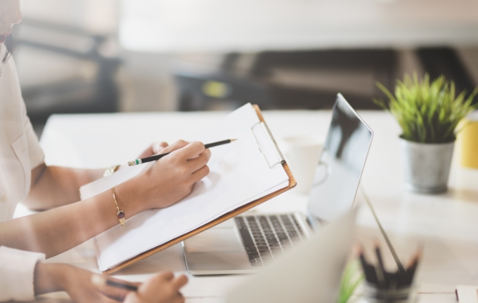 クリップボードにメモを取りながらパソコンを前に作業する人物。求人票の作成や内容検討を示すイメージ